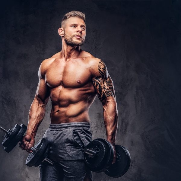 Powerful stylish bodybuilder with tattoo on his arm, doing the exercises with dumbbells. Isolated on a dark background.