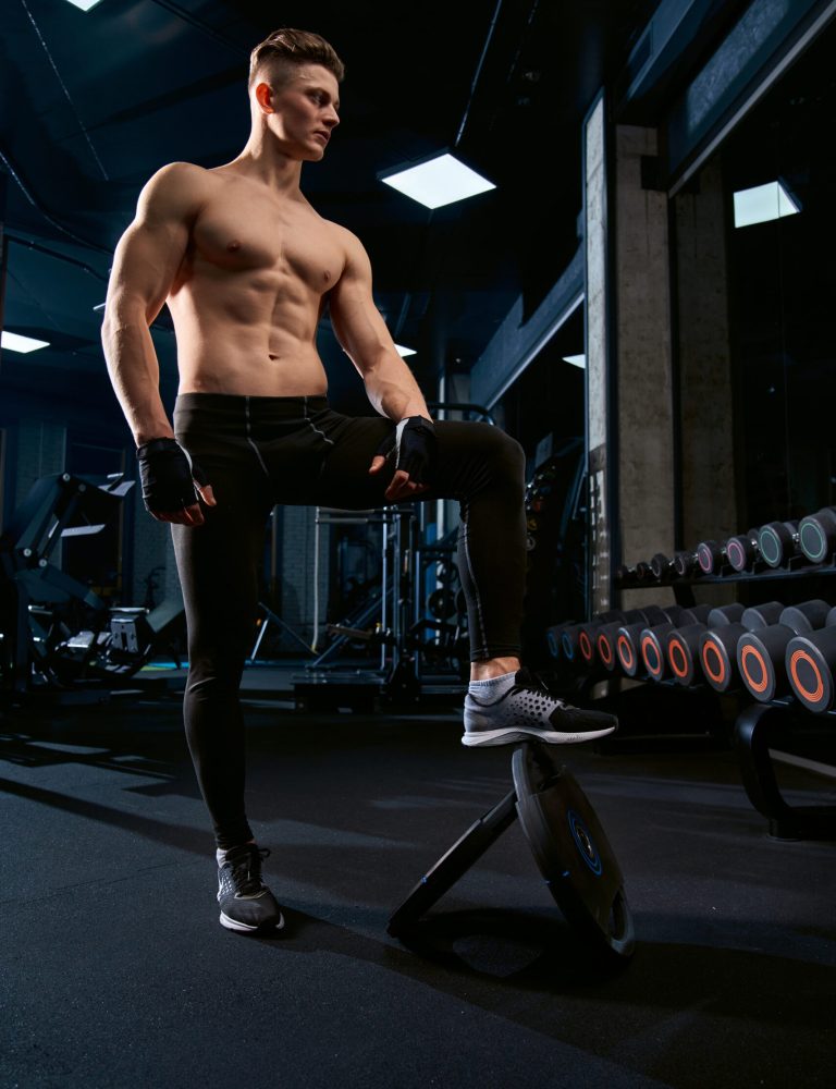 From below of shirtless bodybuilder holding leg on bench. Full length portrait of muscular handsome man with perfect body posing in gym in dark atmosphere. Concept of bodybuilding, healthy lifestyle.